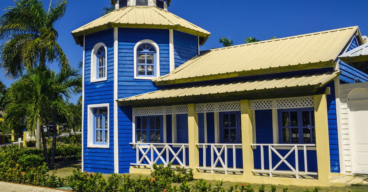 A blue wooden house with a light yellow roof and white door. The house is next to palm trees and some plants.