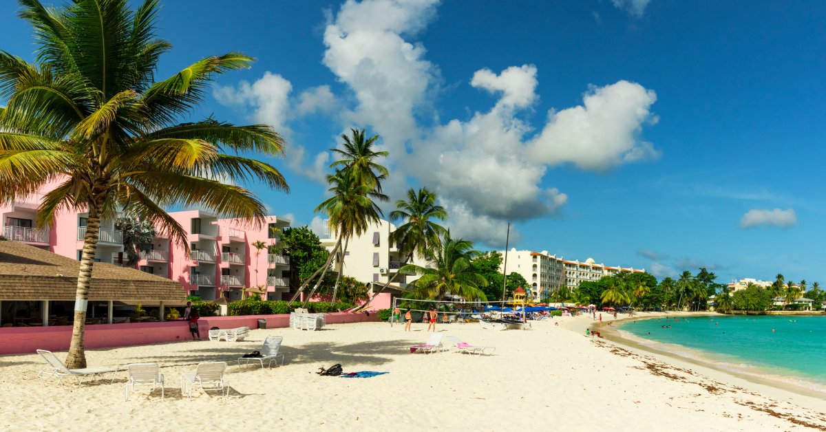 A sandy beach with chairs on it and people playing volleyball. There are properties and palm trees nearby.