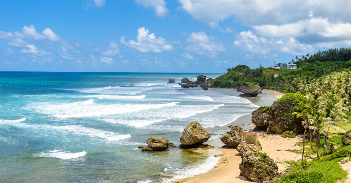 A beach with large rocks and multiple palm trees. There are waves in the ocean and a house in the distance.