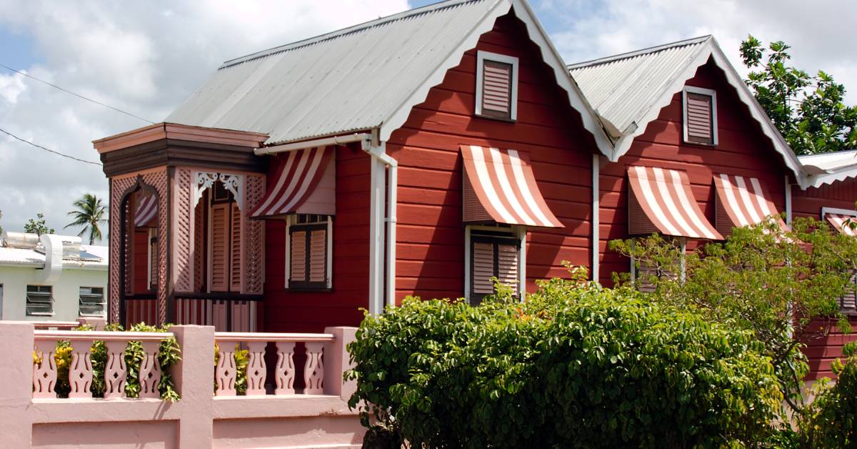 A red house lined with bushes and small trees. The surrounding fence and accents on the house are pink.