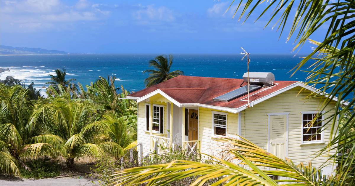 A yellow house that is near multiple green plants. There is a view of the ocean in the distance.