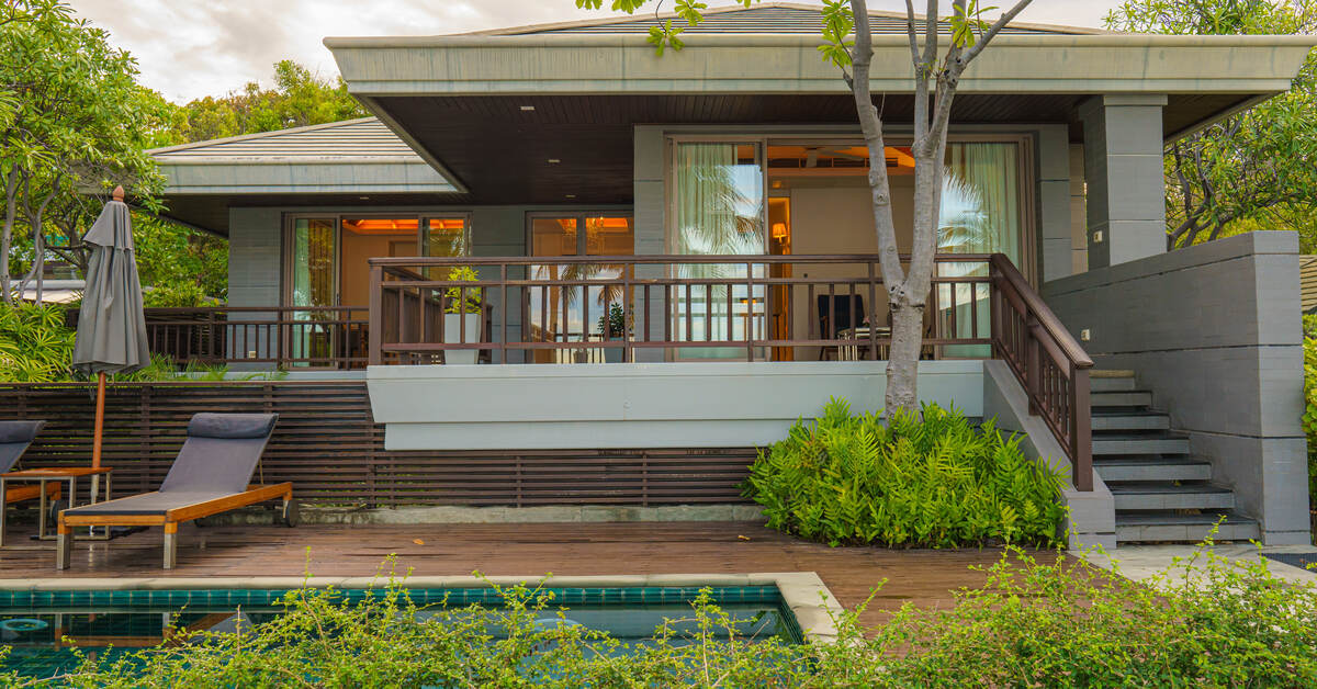 The outside of a modern house with a pool and a deck. Various plants and trees surrounding the house.