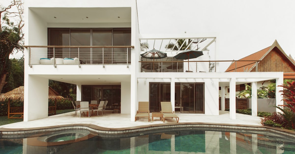 A large white villa with a pool. The villa has two floors and there are multiple outdoor chairs on each level.