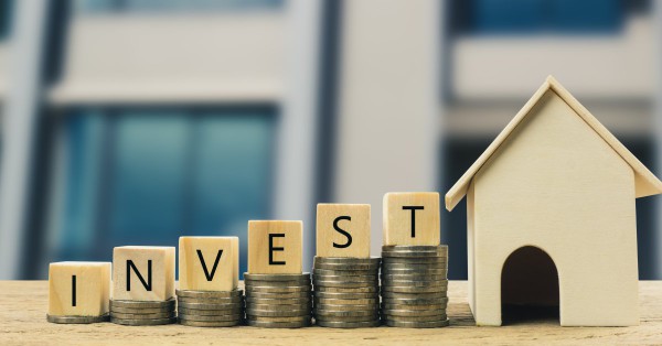 Closeup of a miniature wood house next to building blocks that say invest and are sitting on top of piles of coins.