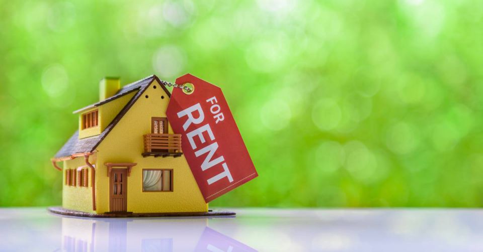 Close-up of a small yellow model of a house with a red 'for rent' tag attached to it in front of a blurred green background.